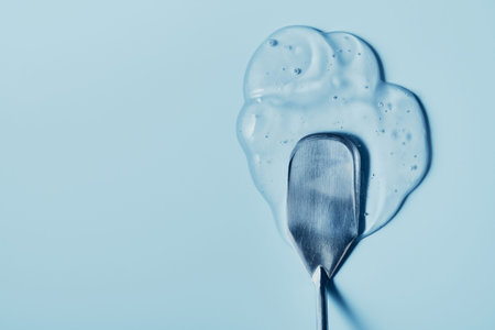 Transparent cosmetic gel spreading with metal spatula on light blue surface creating smooth texture