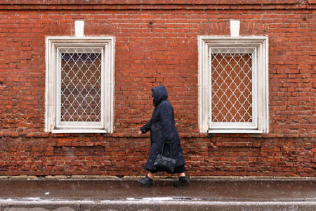 Townspeople. A lonely woman in snowy weather against a brick wall background with two windows. Snowy weather. Woman goes about her business. The townspeople are in a bad weather. Woman is hurrying about her business.の写真素材