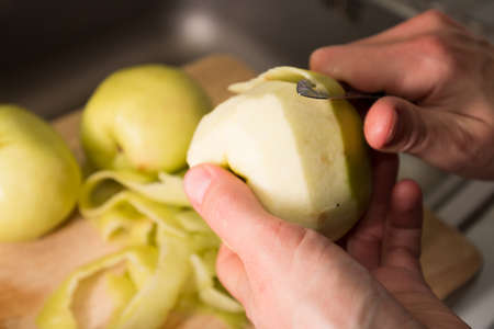 Female hands peeling skin off of green apple using a paring knife. Peel the Skin Off Apples. Woman cuts off the peel of an apple. Garden apples. Golden apples. Farm products. Juicy fruit.の写真素材