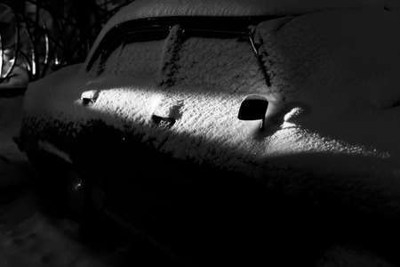 Snow covered old classic car illuminated by a ray of light at night. Old car covered with snow. Soviet car Volga. Details of a car covered with snowの写真素材