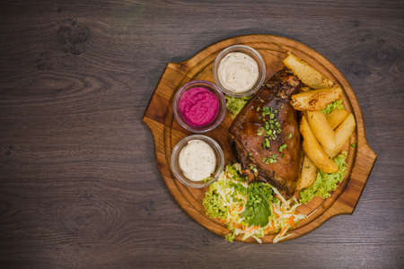 Fried pork leg drumstick, knee of a wild boar with vegetables of red and yellow tomatoes, garlic, parsley and basil and potatoes on a wooden background.の写真素材