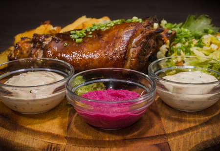 Fried pork leg drumstick, knee of a wild boar with vegetables of red and yellow tomatoes, garlic, parsley and basil and potatoes on a wooden background.の写真素材