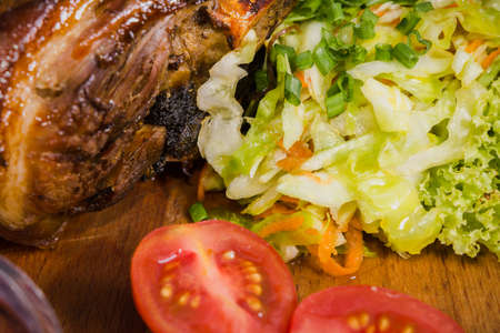 Fried pork leg drumstick, knee of a wild boar with vegetables of red and yellow tomatoes, garlic, parsley and basil and potatoes on a wooden background.の写真素材