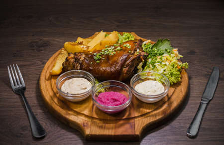Fried pork leg drumstick, knee of a wild boar with vegetables of red and yellow tomatoes, garlic, parsley and basil and potatoes on a wooden background.の写真素材