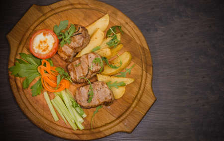 Three pork steaks with fried potatoes and vegetables on a wooden background.の写真素材
