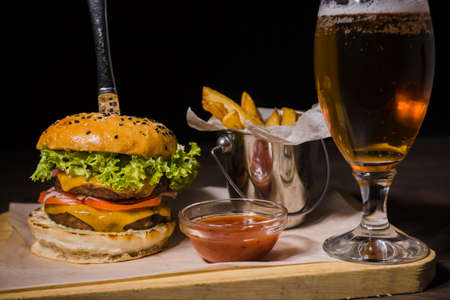Burger with fried potatoes and tomato sauce on a wooden stand with a glass of beer.の写真素材