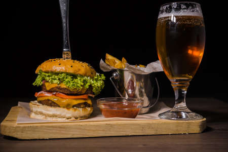 Burger with fried potatoes and tomato sauce on a wooden stand with a glass of beer.の写真素材