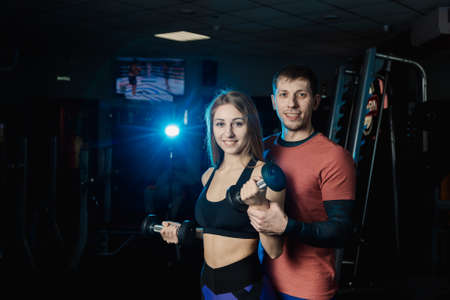 Active beautiful fitness model girl trains biceps with dumbbells together with a trainer in the gym.の写真素材