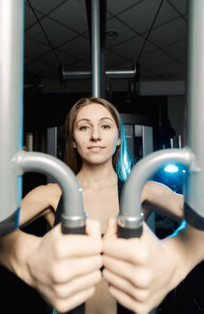 Active beautiful fitness model girl trains pectoral muscles on a simulator in the gym.