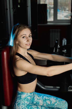 Active beautiful fitness model girl trains pectoral muscles on a simulator in the gym.