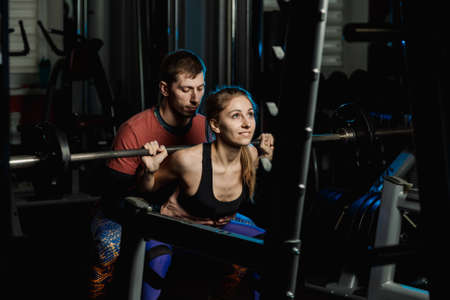 Handsome athletic man helps a beautiful girl to squat with a barbell on the shoulders in the gym.