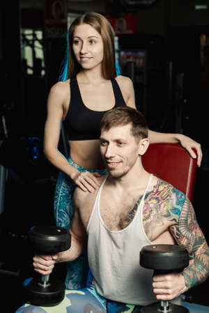 Active beautiful fitness model girl helps to train with dumbbells while sitting to her boyfriend in the gym.の写真素材