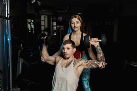 Active beautiful fitness model girl helps to train with dumbbells while sitting to her boyfriend in...