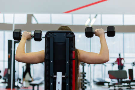 Very beautiful girl trains shoulders with dumbbells on the bench in the gym.の写真素材