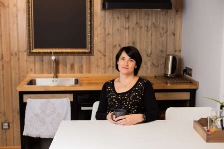 A beautiful woman of 50 years old is sitting in the kitchen in a modern interior and drinking tea. Woman portrait.の写真素材