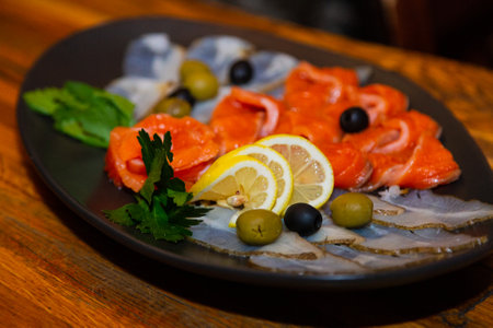 Sliced two varieties of fish with greens, lemon and olives in a plate on a wooden table in a restaurant. large side viewの写真素材