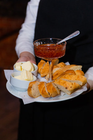 A glass bowl with red jam on a tray with bread and butter in the waiters hands