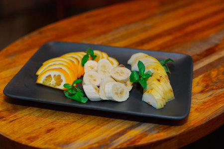 Beautifully chopped fruits on a black plate decorated with mint, all this stands on a wooden tableの写真素材