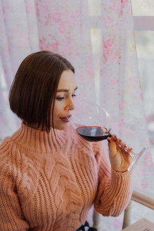 Beautiful young girl sitting at a table in a restaurant with a glass of red wine and posingの写真素材