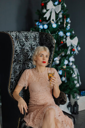 A woman sits on a chair near a Christmas tree with a glass of champagne