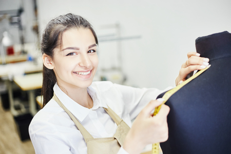 Pretty young woman smiling and looking at camera while measuring shoulders of dummy with tape in workshopの写真素材