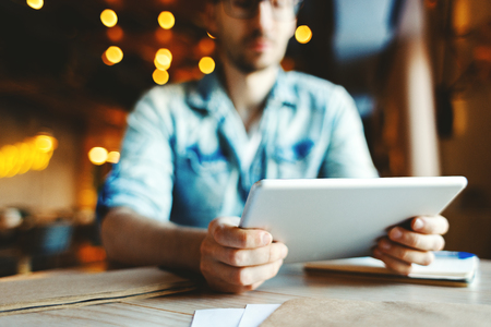 Defocused man sitting at cafe table and holding digital tablet. Guy using tablet computer for working or studyingの写真素材