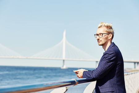 Side view of handsome young blond male in glasses and checkered jacket standing relaxed at handrail with closed eyes on blurred background of water surface and suspension bridgeの写真素材