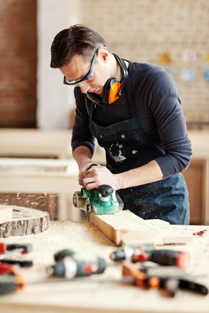 Young woodworker sanding wooden plank with flatbed sander machine, his workbench covered with tools, wood shavings and sawdustの写真素材
