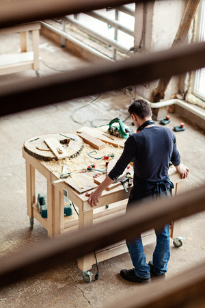 From above back through fence view of young woodworker looking at wooden details at workbenchの写真素材
