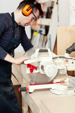 Side view of young woodworker in protective eyeglasses and ear muffs sawing wooden plank with electric circular sawの写真素材