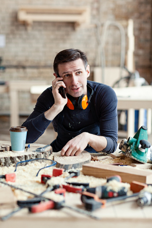 Young woodworker talking on mobile phone at workbench surrounded by tools and wood slicesの写真素材