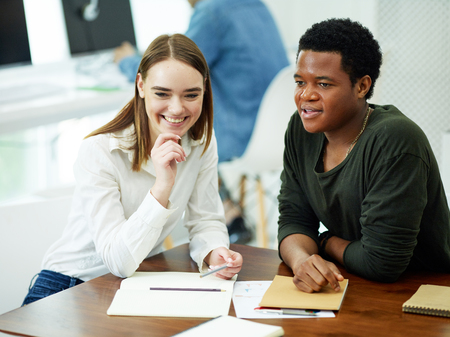 Caucasian young woman sitting at table with African-American man with notebooks smiling and looking one way in university libraryの写真素材