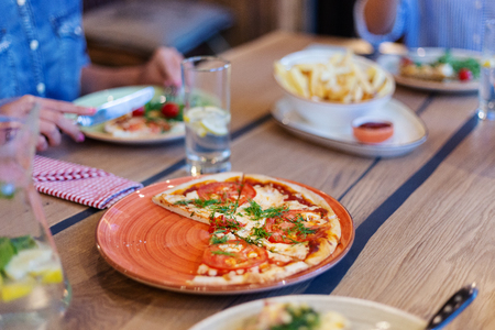 Slices of pizza on red plate on wooden dining table surrounded by other food and drinks and defocused people dining in backgroundの写真素材