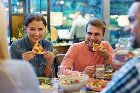 Cheerful young people enjoying eating delicious pizza and talking during dinner with friends in restaurantの写真素材