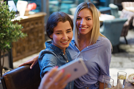 Two smiling young woman posing together for selfie on smartphone at table in restaurantの写真素材