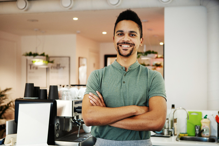 Happy ethnic bartender with crossed arms looking at camera while standing at counter in coffee shopの写真素材