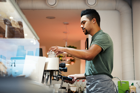Side view of young ethnic bartender pouring coffee into cup from coffee machine at counter in coffee shopの写真素材
