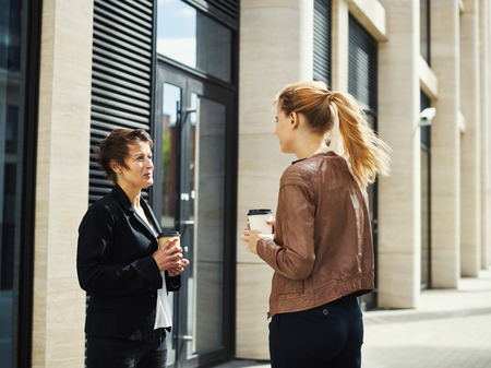 Middle-aged and young women in formal outfits having cup of coffee and talking in leisure outside on streetの写真素材