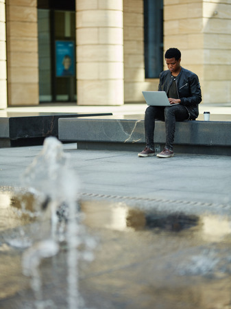 Young African American entrepreneur siting on stone seat near fountain outdoors and working online on his laptopの写真素材