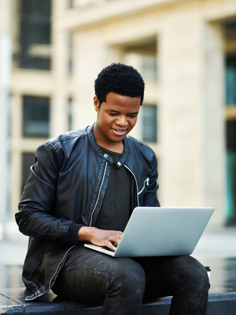 African young man smiling while browsing social media on laptop computer outsideの写真素材