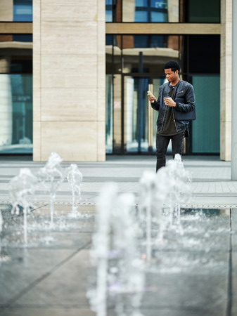 African American young man standing by fountain in city center and checking text messages on his cell phone, paper coffee cup in his hand and laptop under armの写真素材