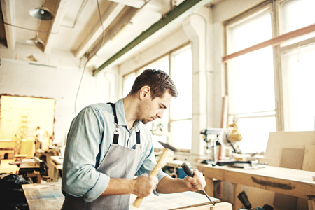 Young carpenter working with timber in his shop, using hammer and wood chiselの写真素材