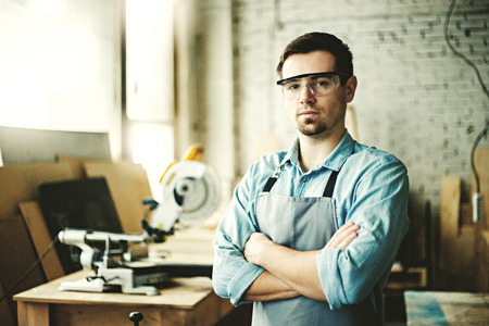 Portrait of young craftsman in protective eyewear posing cross-armed in his own woodworking shop, copy space to the leftの写真素材