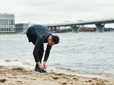 Side view of young guy in sportswear standing on sand near waving water and tying laces on sneakersの写真素材