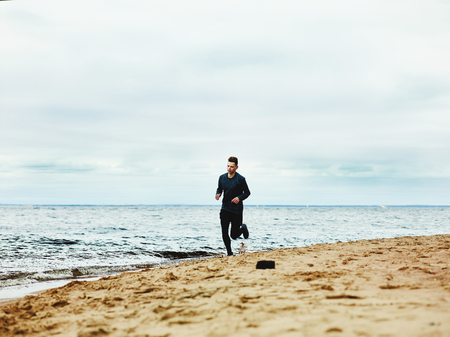 Young man in sportswear running on sandy coast near water while training on dull overcast dayの写真素材
