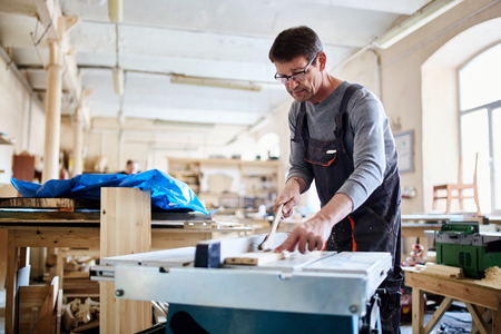 Adult man in glasses in workshop creating furniture of wood with table sawの写真素材