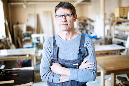 Adult man in overall and eyeglasses holding hands crossed and looking at camera in modern joineryの写真素材
