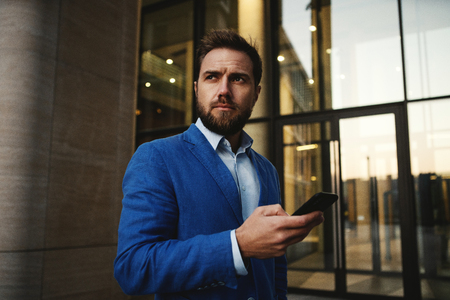 Contemporary stylish man wearing suit and using cellphone on street with background of modern building looking awayの写真素材
