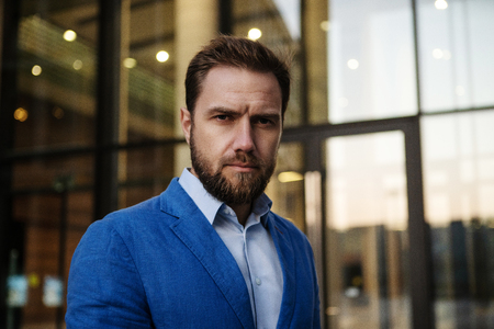 Handsome adult businessman with beard wearing blue suit and looking seriously at camera outdoorsの写真素材