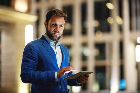 Modern stylish man in suit holding tablet and looking seriously at camera on blurred background of night buildings in lightsの写真素材